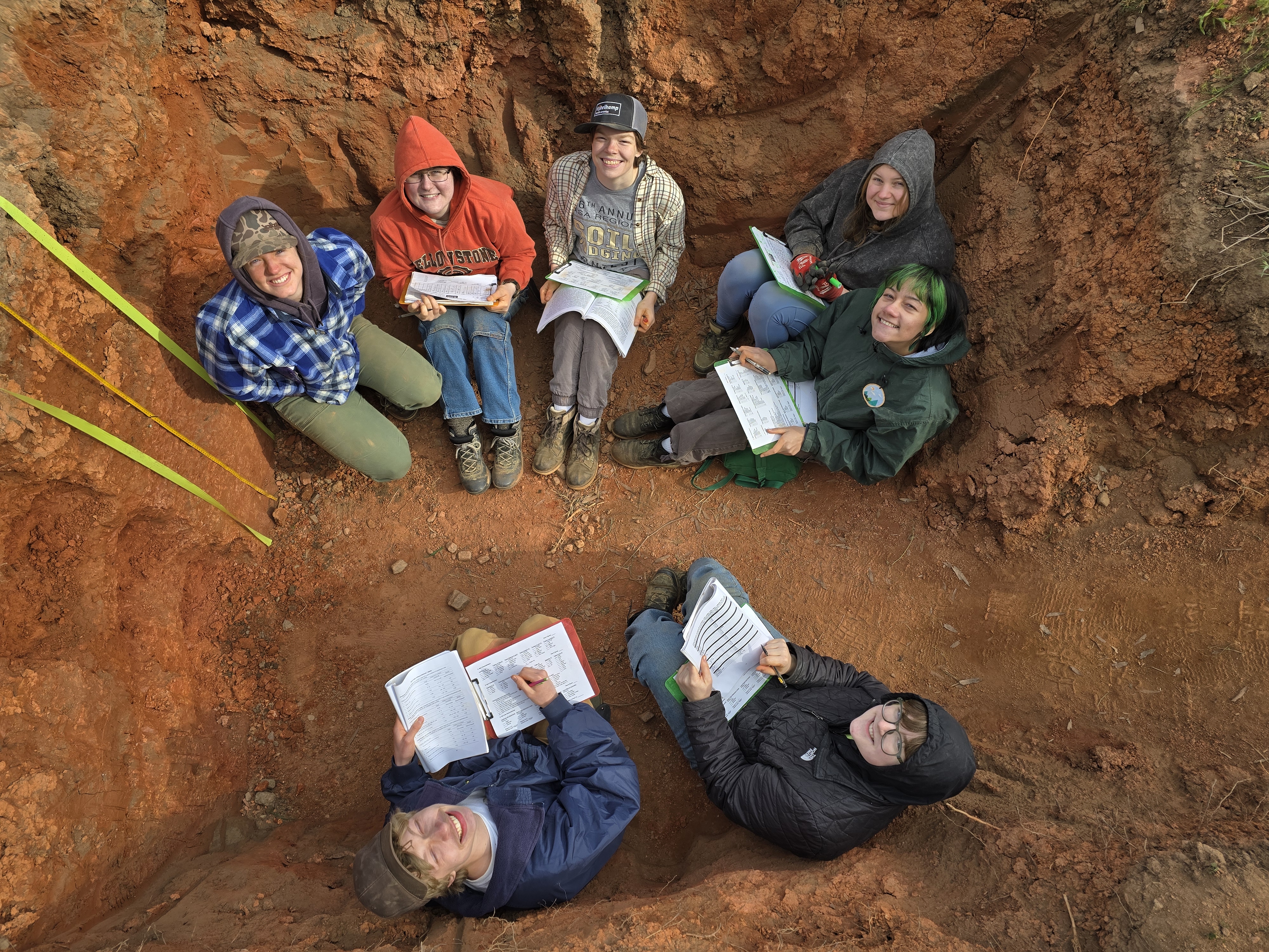 Members of the MSU Soil Judging Team sit in a circle in a pit and look up smiling at the photographer. Several of them are holding notebooks.